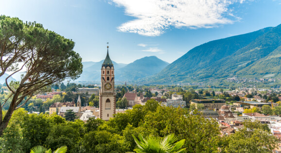 Panoramablick über Meran mit mediterraner Vegetation und den Südtiroler Bergen im Hintergrund.