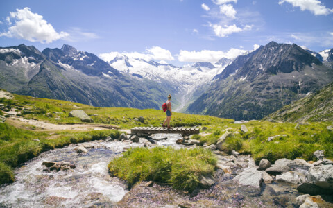 Wanderer im Zillertal überquert einen Gebirgsbach mit Blick auf schneebedeckte Alpen.