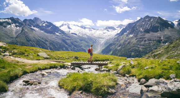 Wanderer im Zillertal überquert einen Gebirgsbach mit Blick auf schneebedeckte Alpen.